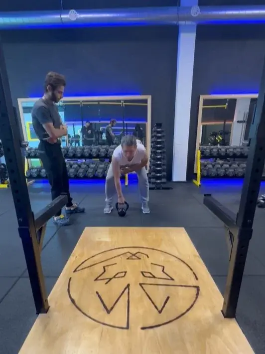 Een coach observeert een klant bij een kettlebell-oefening op het platform met het wolflogo